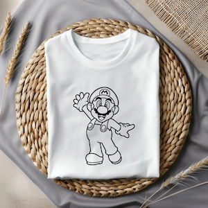 May include: White t-shirt featuring a black outline illustration of the video game character Mario waving. The character is wearing his signature hat, overalls, and gloves. The shirt is folded on a woven, circular surface.