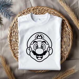 May include: White t-shirt featuring a black outline graphic of Mario's face. The cartoon character has a large mustache, a hat with an "M" symbol, and a smiling expression. The shirt is displayed on a woven, circular placemat.