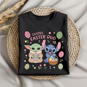 Könnte beinhalten: Schwarzes T-Shirt mit einem Motiv von Baby Yoda und Stitch, die Ostereier halten. Der Text "Cutes Easter Duo" ist auf dem T-Shirt aufgedruckt.