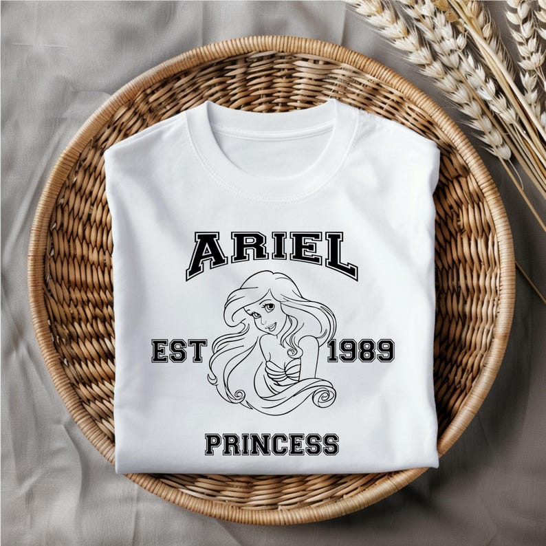Puede incluir: Camiseta blanca con un gr&aacute;fico negro de Ariel de La Sirenita. El gr&aacute;fico incluye el texto "Ariel Est 1989 Princess".