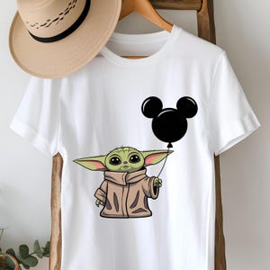 K&ouml;nnte beinhalten: Wei&szlig;es T-Shirt mit einer gr&uuml;nen Figur, die einen schwarzen Mickey Mouse-Ballon h&auml;lt.
