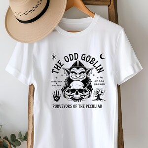 May include: White t-shirt with a black and white graphic design of a goblin holding a skull. The text "The Odd Goblin" is written in a circular design around the goblin. The text "Oddities & Curios of the Peculiar" is written below the goblin.