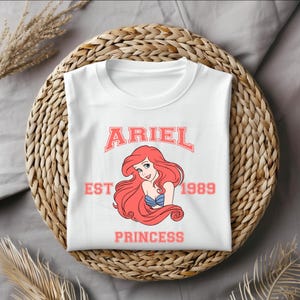 Puede incluir: Camiseta blanca con un gr&aacute;fico rosa de Ariel de La Sirenita. El gr&aacute;fico incluye el texto "Ariel Est 1989 Princess".