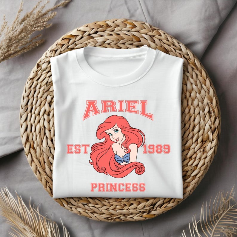 Puede incluir: Camiseta blanca con un gr&aacute;fico rosa de Ariel de La Sirenita. El gr&aacute;fico incluye el texto "Ariel Est 1989 Princess".