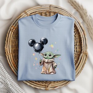 Op de afbeelding: Een lichtblauw T-shirt met een cartoon afbeelding van een groene alien die een zwarte Mickey Mouse ballon vasthoudt. De alien draagt een bruine robe en heeft grote oren. De achtergrond is lichtblauw met witte sterren.