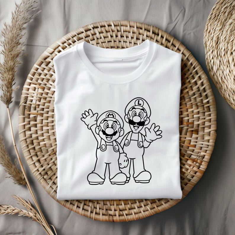 May include: White t-shirt featuring a black outline illustration of two cartoon characters, one with an "M" on their hat and the other with an "L". Both characters are waving. The shirt is displayed on a woven basket.
