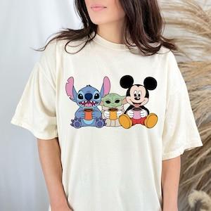 Puede incluir: Una camiseta blanca con un gr&aacute;fico de Stitch, Baby Yoda y Mickey Mouse sosteniendo tazas. Los personajes son de estilo de dibujos animados y tienen una expresi&oacute;n amigable.