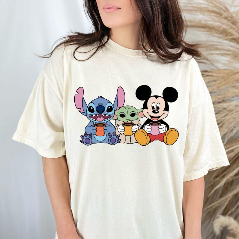 Puede incluir: Una camiseta blanca con un gr&aacute;fico de Stitch, Baby Yoda y Mickey Mouse sosteniendo tazas. Los personajes son de estilo de dibujos animados y tienen una expresi&oacute;n amigable.
