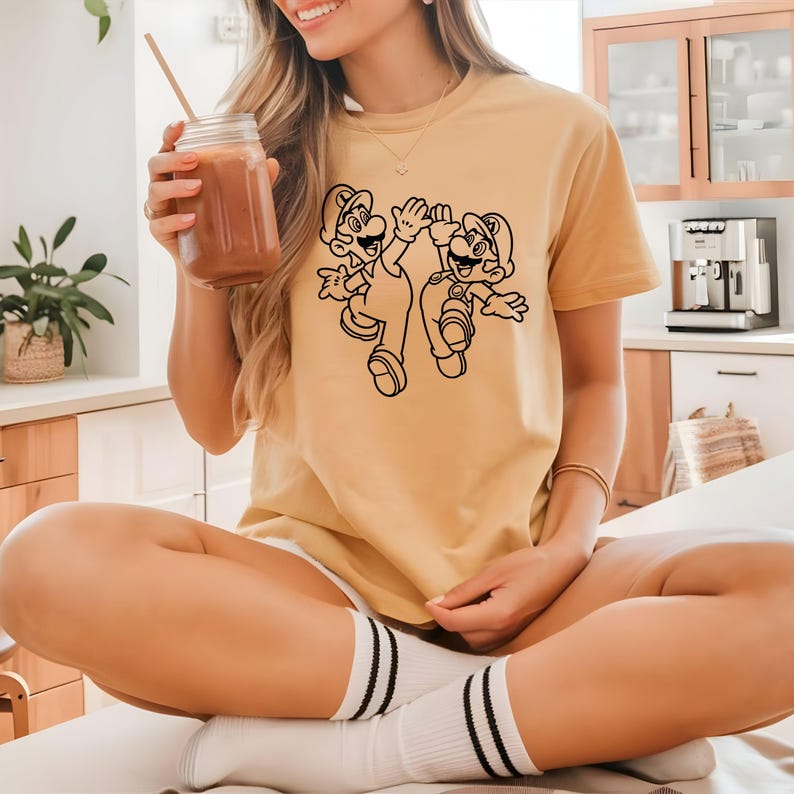 May include: Mustard yellow t-shirt featuring a black outline graphic of two cartoon characters. The characters are wearing overalls and hats, with arms raised in a celebratory gesture. The shirt is short-sleeved and appears to be made of soft material.
