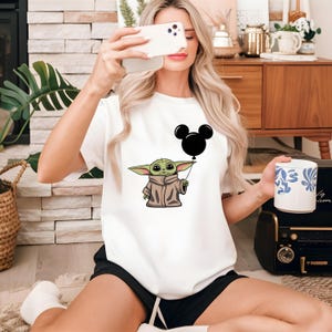 K&ouml;nnte beinhalten: Wei&szlig;es T-Shirt mit einer gr&uuml;nen Comicfigur, die einen schwarzen Mickey-Maus-Ballon h&auml;lt.