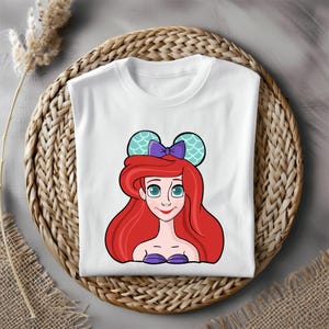Puede incluir: Camiseta blanca con una imagen de dibujos animados de Ariel de La Sirenita con un lazo morado y orejas de rat&oacute;n de color turquesa con un patr&oacute;n de escamas.