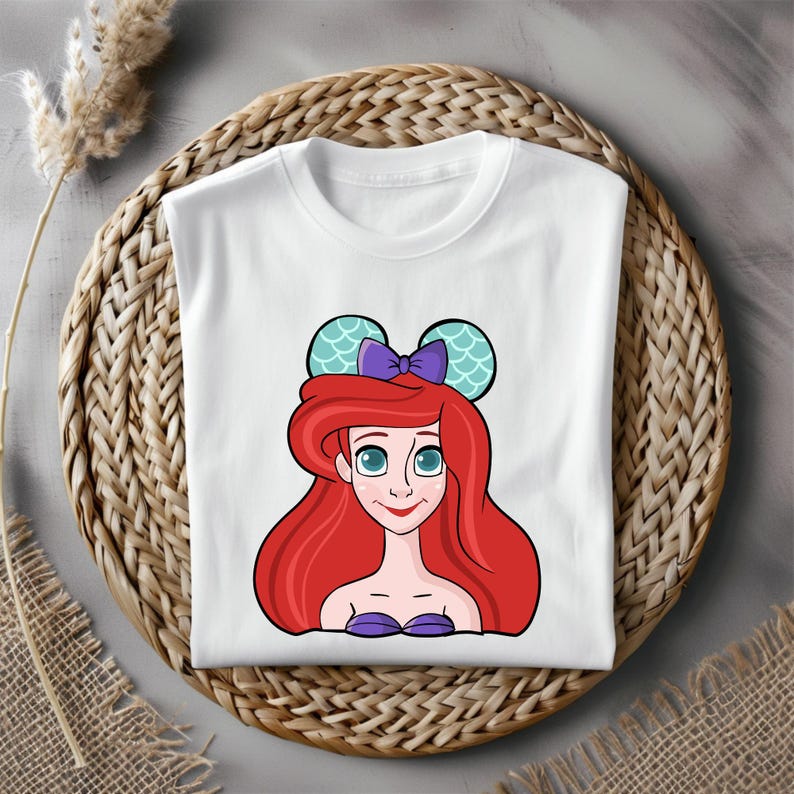 Puede incluir: Camiseta blanca con una imagen de dibujos animados de Ariel de La Sirenita con un lazo morado y orejas de rat&oacute;n de color turquesa con un patr&oacute;n de escamas.