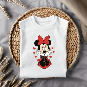 May include: White t-shirt with a red and black cartoon image of Minnie Mouse. Minnie Mouse is wearing a red dress and a red bow. She is surrounded by red hearts.