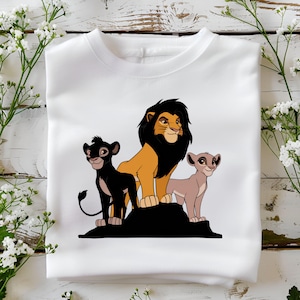 May include: White t-shirt with a graphic of three lions from the Lion King movie. The lions are standing on a rock. The lion in the middle is a large, black-maned lion with a golden body. The lion on the left is a black lion with a dark mane. The lion on the right is a light brown lion with a light brown mane.