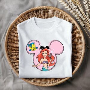 Puede incluir: Camiseta blanca con un dise&ntilde;o de orejas de Mickey Mouse rosa con Ariel de La Sirenita sosteniendo un helado de Mickey Mouse y Flounder nadando a su lado.