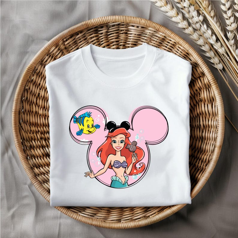 Puede incluir: Camiseta blanca con un dise&ntilde;o de orejas de Mickey Mouse rosa con Ariel de La Sirenita sosteniendo un helado de Mickey Mouse y Flounder nadando a su lado.