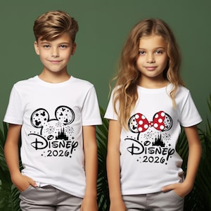 May include: Two white t-shirts with black Disney-themed graphics. One shirt features Mickey Mouse ears with stars, fireworks, and the text "Disney 2026!". The other shirt has Minnie Mouse ears with a red polka-dot bow and the same text.