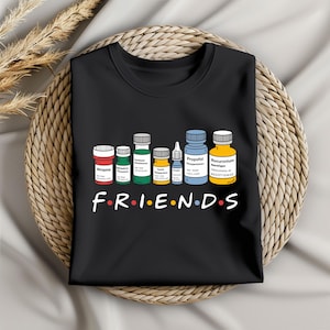 May include: Black t-shirt with a line of colorful medicine bottles and the word "FRIENDS" written below. Each bottle has a different name of a medication printed on the label.