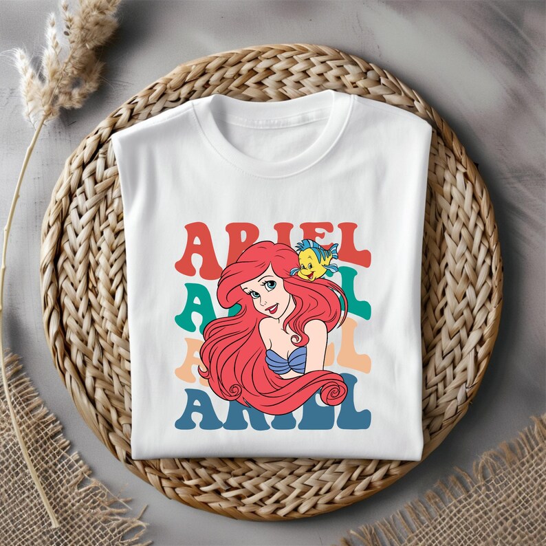 Puede incluir: Camiseta blanca con un gr&aacute;fico retro de Ariel de La Sirenita. El gr&aacute;fico presenta a Ariel con su cabello rojo y un pez amarillo, con la palabra "Ariel" repetida en texto colorido y superpuesto.