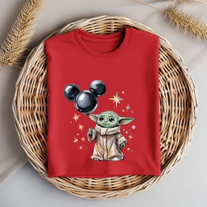 Op de afbeelding: Rood T-shirt met een groen personage dat een zwarte Mickey Mouse ballon vasthoudt. Het personage is omringd door kleine, kleurrijke sterren.
