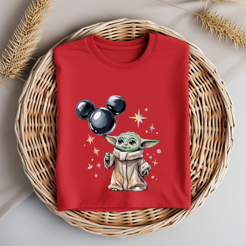 Op de afbeelding: Rood T-shirt met een groen personage dat een zwarte Mickey Mouse ballon vasthoudt. Het personage is omringd door kleine, kleurrijke sterren.