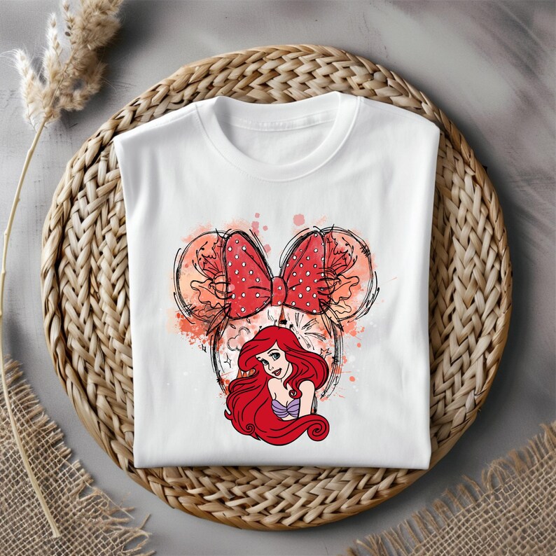 Puede incluir: Camiseta blanca con un lazo rojo y blanco y una imagen de dibujos animados de Ariel de La Sirenita. El lazo tiene la forma de las orejas de Mickey Mouse.