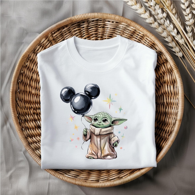 Op de afbeelding: Wit T-shirt met een groen alienfiguur dat een zwarte Mickey Mouse-ballon vasthoudt. Het alien draagt een bruine robe en heeft grote oren. De achtergrond is wit met kleurrijke sterren.