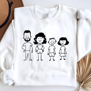 May include: White crewneck sweatshirt featuring a black line drawing of a family of four. The illustration includes a man with a beard, a woman, and two children. The design is simple and cartoon-like.