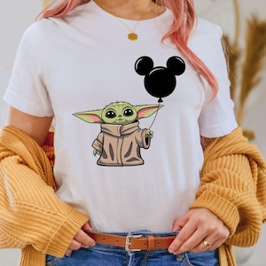 K&ouml;nnte beinhalten: Wei&szlig;es T-Shirt mit einer gr&uuml;nen Comicfigur, die einen schwarzen Mickey-Maus-Ballon h&auml;lt.