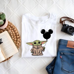 K&ouml;nnte beinhalten: Wei&szlig;es T-Shirt mit einer gr&uuml;nen Comicfigur, die einen schwarzen Mickey-Maus-Ballon h&auml;lt. Die Figur tr&auml;gt einen braunen Mantel.