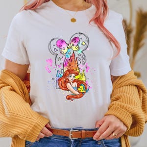 Puede incluir: Camiseta blanca con una ilustraci&oacute;n colorida de Ariel de La Sirenita sosteniendo a Flounder. La ilustraci&oacute;n incluye un castillo y una silueta de oreja de Mickey Mouse.