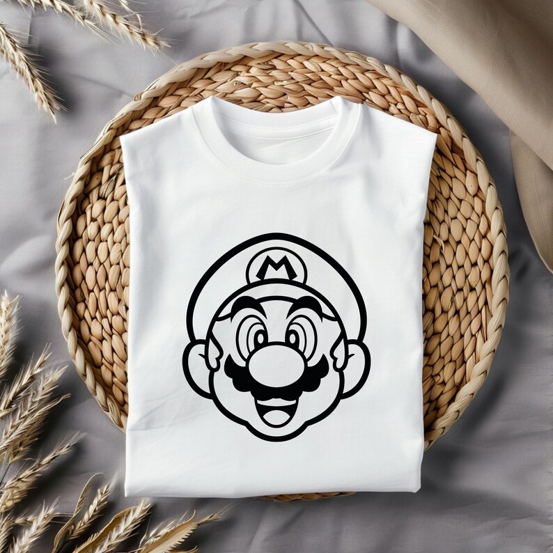 May include: White t-shirt featuring a black graphic of Mario's face. The design includes his hat with the letter "M", a mustache, and a smiling expression. The shirt is folded on a woven basket.
