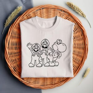 May include: White t-shirt featuring a black outline graphic of Mario, Luigi, and Yoshi. The characters are waving and standing side-by-side. The shirt is folded and placed on a woven wicker basket.