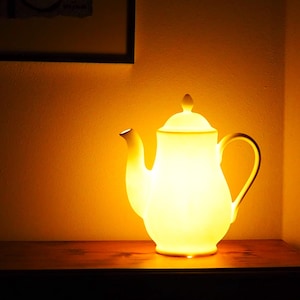 May include: A glowing white teapot-shaped lamp with a curved handle and spout. The lamp is illuminated from within, casting a warm, yellow light. It sits on a dark wooden surface against a yellow wall.