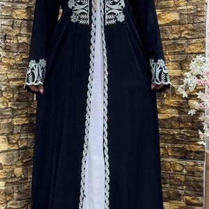 May include: A black abaya with white embroidery. The abaya has long sleeves and a long, flowing skirt. The embroidery is intricate and detailed, and it covers the entire front of the abaya. The abaya is open in the front, and it is worn over a white dress.