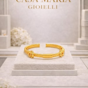 Puede incluir: Un brazalete dorado con un diseño abierto, exhibido sobre una superficie blanca. El brazalete presenta detalles intrincados y un patrón texturizado. La marca "CASA MARIA GIOIELLI" es visible en el fondo.