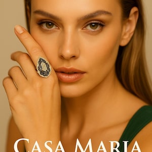 May include: A close-up of a woman's face with a large, teardrop-shaped ring. The ring features a black and gold design with small, sparkling stones. The text "CASA MARIA GIOIELLI" is displayed in white.