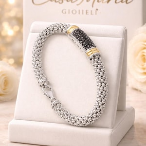 May include: A silver bracelet with a textured design and a black stone center, accented with gold. The bracelet is displayed on a white stand, with the "Casa Maria Gioielli" logo in the background.