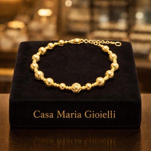 Op de afbeelding: Een gouden kralenarmband tentoongesteld op een zwarte fluwelen doos. De armband heeft verschillende maten bolvormige kralen die aan elkaar zijn geregen. De doos heeft de tekst "Casa Maria Gioielli" in gouden letters.
