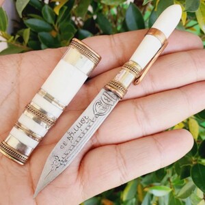 May include: An ornate knife with a white handle and a silver blade. The handle is decorated with gold and silver bands. The knife is in a sheath with matching gold and silver accents. The blade has etched details and text.