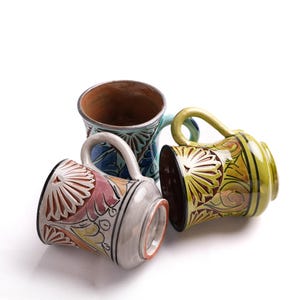 May include: Three ceramic mugs featuring intricate hand-painted floral patterns in vibrant shades of yellow, pink, and blue. Each mug has a sturdy handle and a glossy finish, with one mug standing upright and two positioned on their sides.