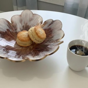 May include: A scalloped, leaf-shaped bowl featuring a swirling white and brown resin pattern with a gold-toned rim, containing two round, golden-brown baked scones. Beside the bowl sits a white ceramic mug filled with steaming black coffee.
