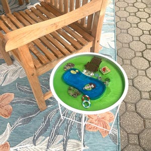 May include: A miniature pool scene on a white-legged side table. The pool is blue with a person floating inside. The surrounding area is green with miniature accessories. A wooden chair sits nearby on a floral rug.