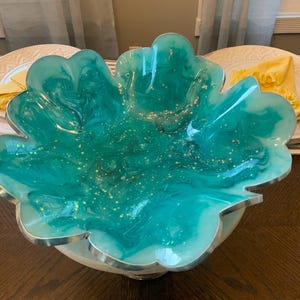 Peut inclure: Un bol décoratif en forme de grande feuille, présentant un mélange tourbillonnant de résine bleu sarcelle translucide et blanche opaque. La surface est incrustée de fines paillettes dorées et les bords sont finis avec une bordure argentée métallique.