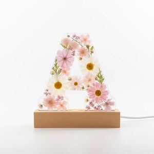 May include: A clear resin letter A filled with pressed pink and white flowers, including daisies and small blossoms. The letter is mounted on a rectangular light-colored wooden base with a power cord extending from the right side of the base.