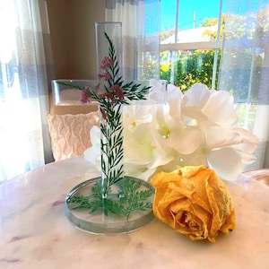 May include: A clear resin cross with embedded greenery and pink flowers, set on a round base. A dried yellow rose and white flowers are next to the cross on a marble-look table. The scene is lit by natural light.