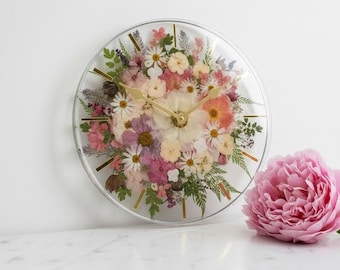 May include: A round wall clock featuring a clear face filled with an arrangement of pressed dried flowers in shades of pink, white, and green. The clock has thin gold-toned hour markers and matching gold-toned hands, accompanied by a large pink peony.