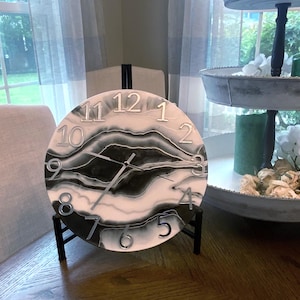 May include: Round wall clock with a black and white abstract design. The clock features silver-colored numerals and hands. It is displayed on a black stand. The clock's face has a marbled appearance, adding a decorative touch to any room.