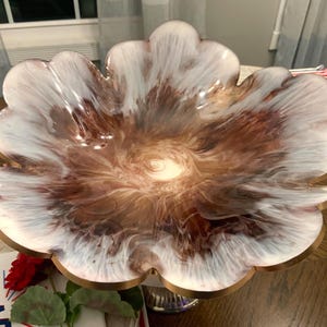 May include: A decorative, flower-shaped bowl with a swirling pattern of white, brown, and gold. The bowl has a gold-colored rim and is elevated on a silver-colored pedestal. The bowl is likely made of resin or a similar material.
