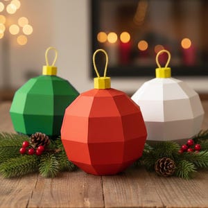 May include: Three geometric Christmas ornaments in green, red, and white, each with a yellow top and loop. They rest on a bed of pine needles, red berries, and pine cones, set against a blurred background of a fireplace and lights.